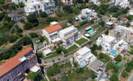 Aerial view of a hillside residential area featuring white houses with orange roofs, green gardens, and a swimming pool, surrounded by lush vegetation.