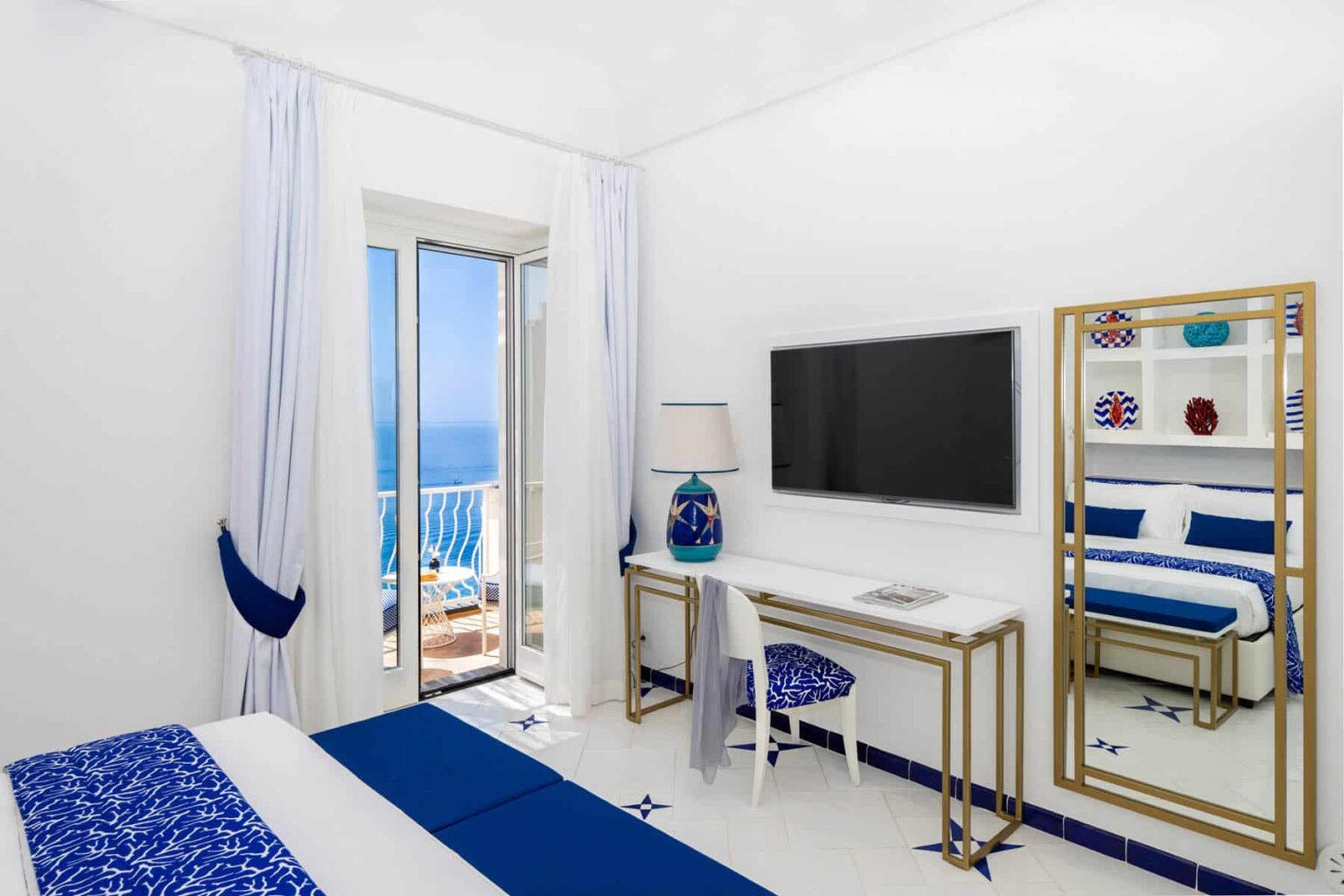 Bright and modern hotel room featuring a blue and white color scheme, a comfortable bed, a desk with a chair, a flat-screen TV, and large windows offering a view of the sea. | Only on SopranoVillas