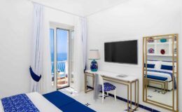 Bright and modern hotel room featuring a blue and white color scheme, a comfortable bed, a desk with a chair, a flat-screen TV, and large windows offering a view of the sea.