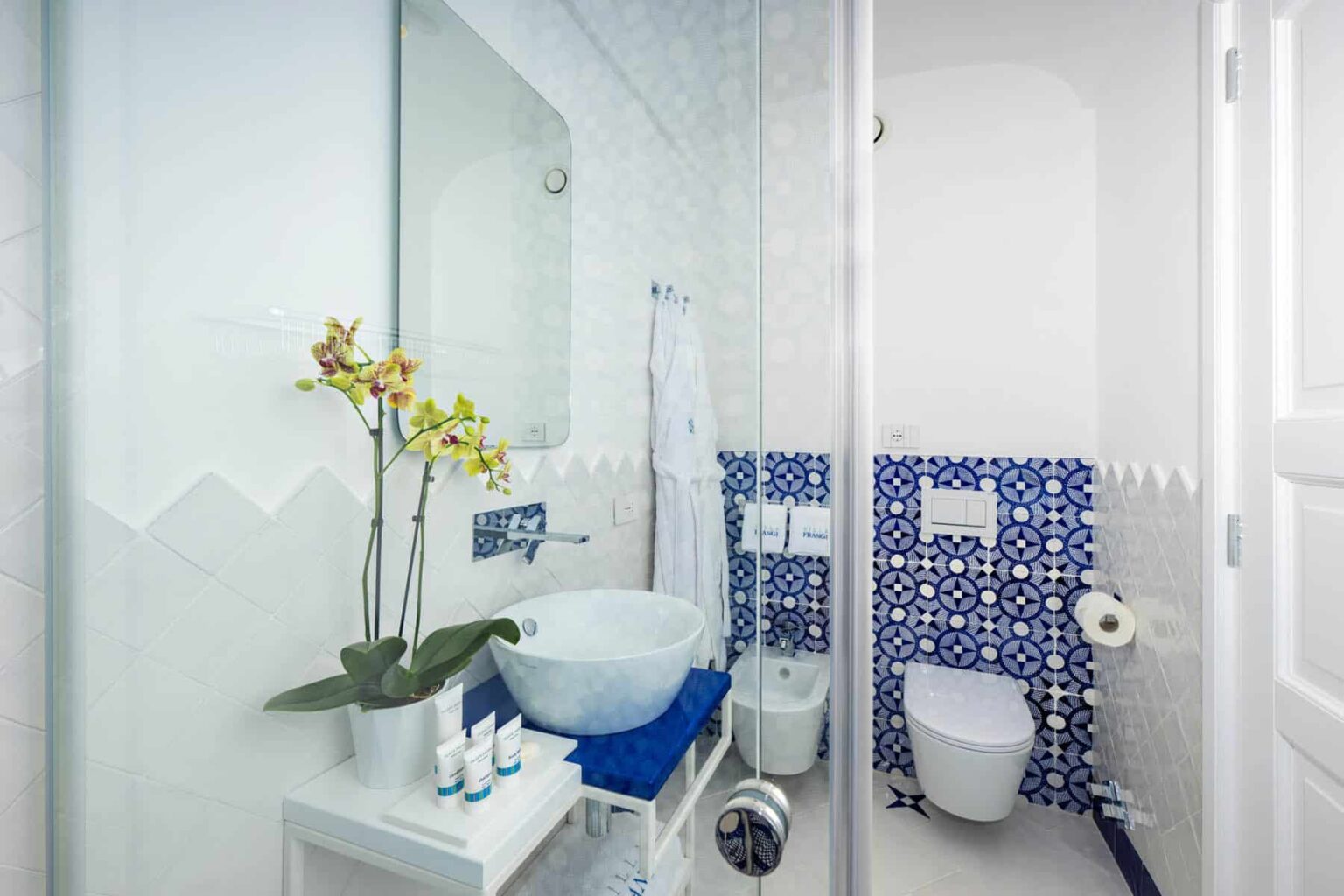 Modern bathroom interior featuring a stylish glass shower, a round basin sink, and decorative blue patterned tiles. The space is enhanced by a touch of greenery with an orchid plant and neatly arranged toiletries, creating a fresh and inviting atmosphere. | Only on SopranoVillas