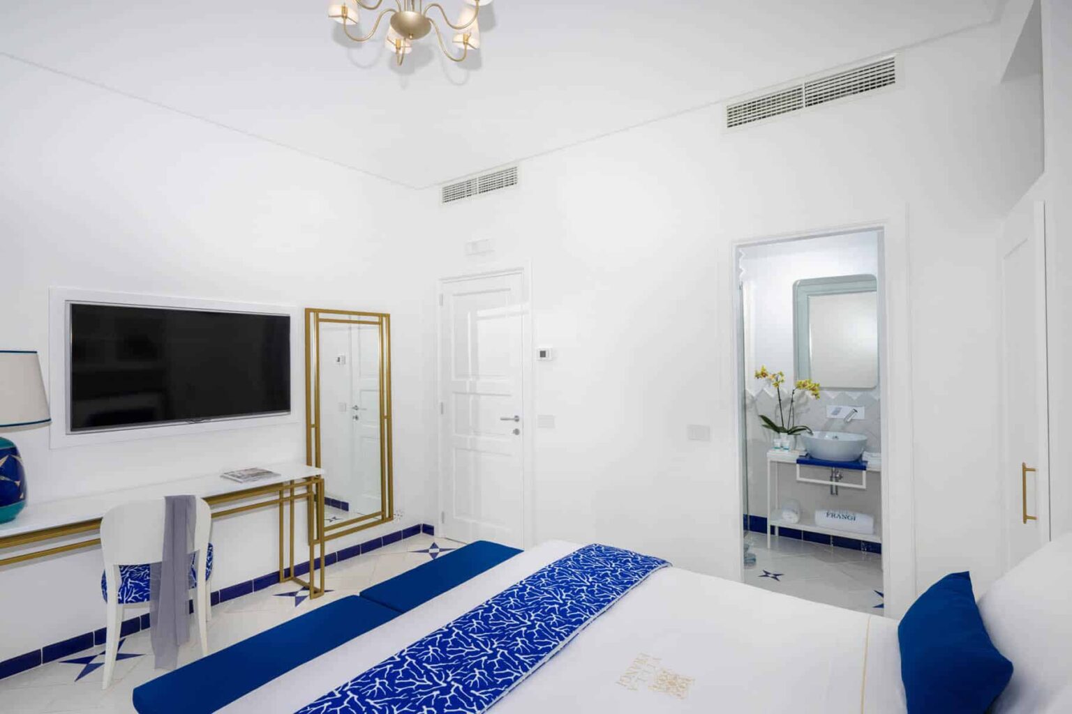 Modern hotel room featuring a white color scheme, a comfortable bed with blue accents, a flat-screen TV, and a stylish bathroom visible through a glass door. Ideal for relaxation and comfort. | Only on SopranoVillas