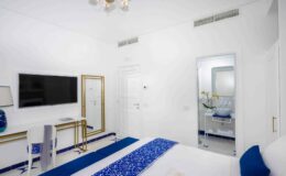 Modern hotel room featuring a white color scheme, a comfortable bed with blue accents, a flat-screen TV, and a stylish bathroom visible through a glass door. Ideal for relaxation and comfort.