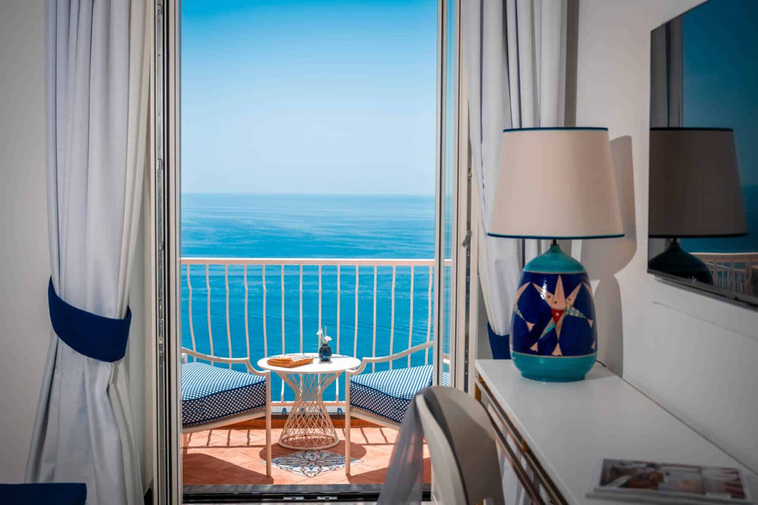 Cozy hotel room with a stunning ocean view, featuring a balcony, a decorative lamp, and a small table with refreshments. Bright curtains frame the scenic landscape, creating an inviting atmosphere. | Only on SopranoVillas