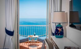 Cozy hotel room with a stunning ocean view, featuring a balcony, a decorative lamp, and a small table with refreshments. Bright curtains frame the scenic landscape, creating an inviting atmosphere.