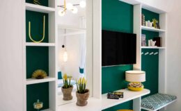 Modern living room interior featuring a green accent wall, white shelving units, a flat-screen TV, decorative plants, and stylish lighting.