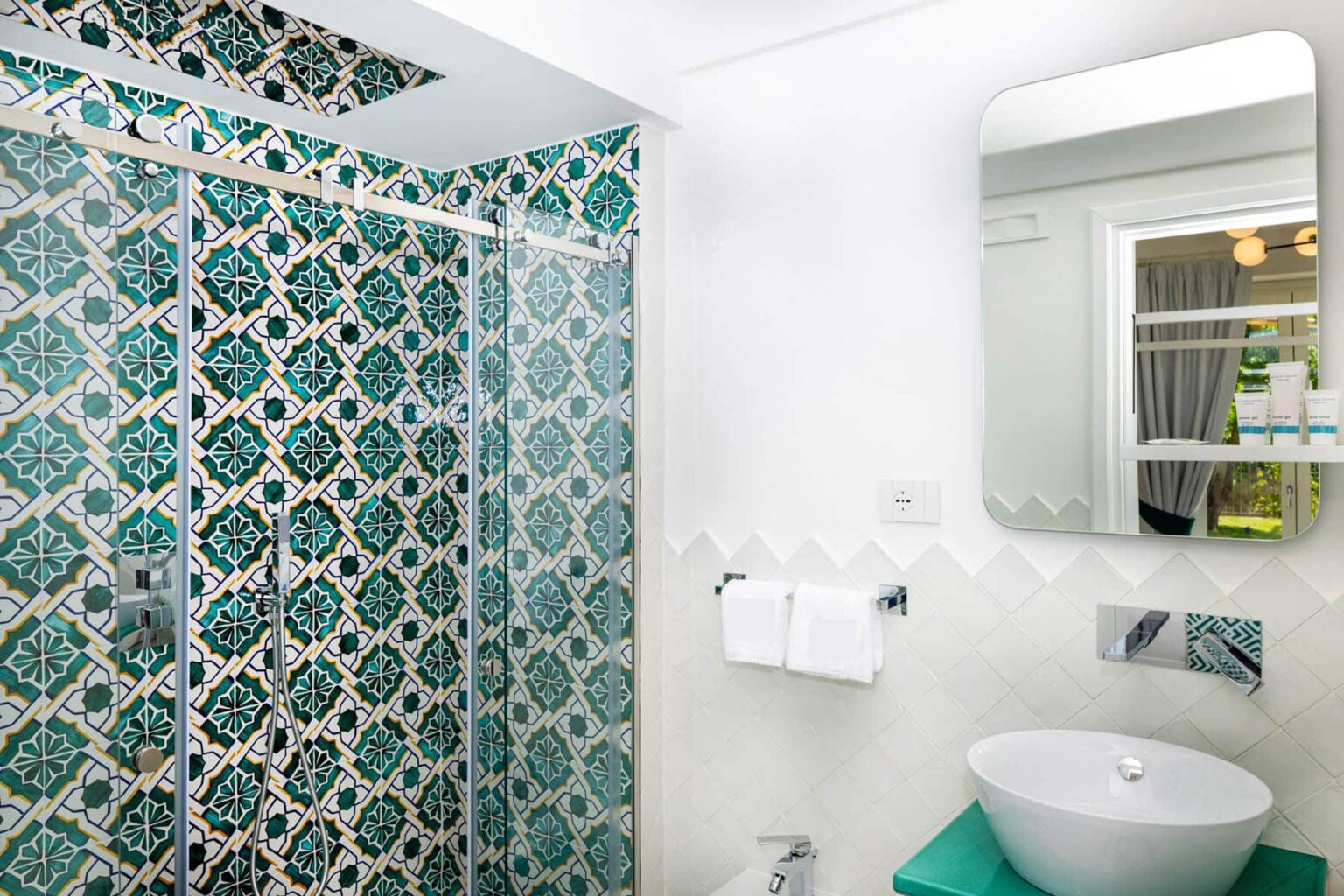Modern bathroom with a stylish glass shower featuring turquoise patterned tiles, a sleek mirror above a contemporary sink, and white towels hanging on the wall. | Only on SopranoVillas