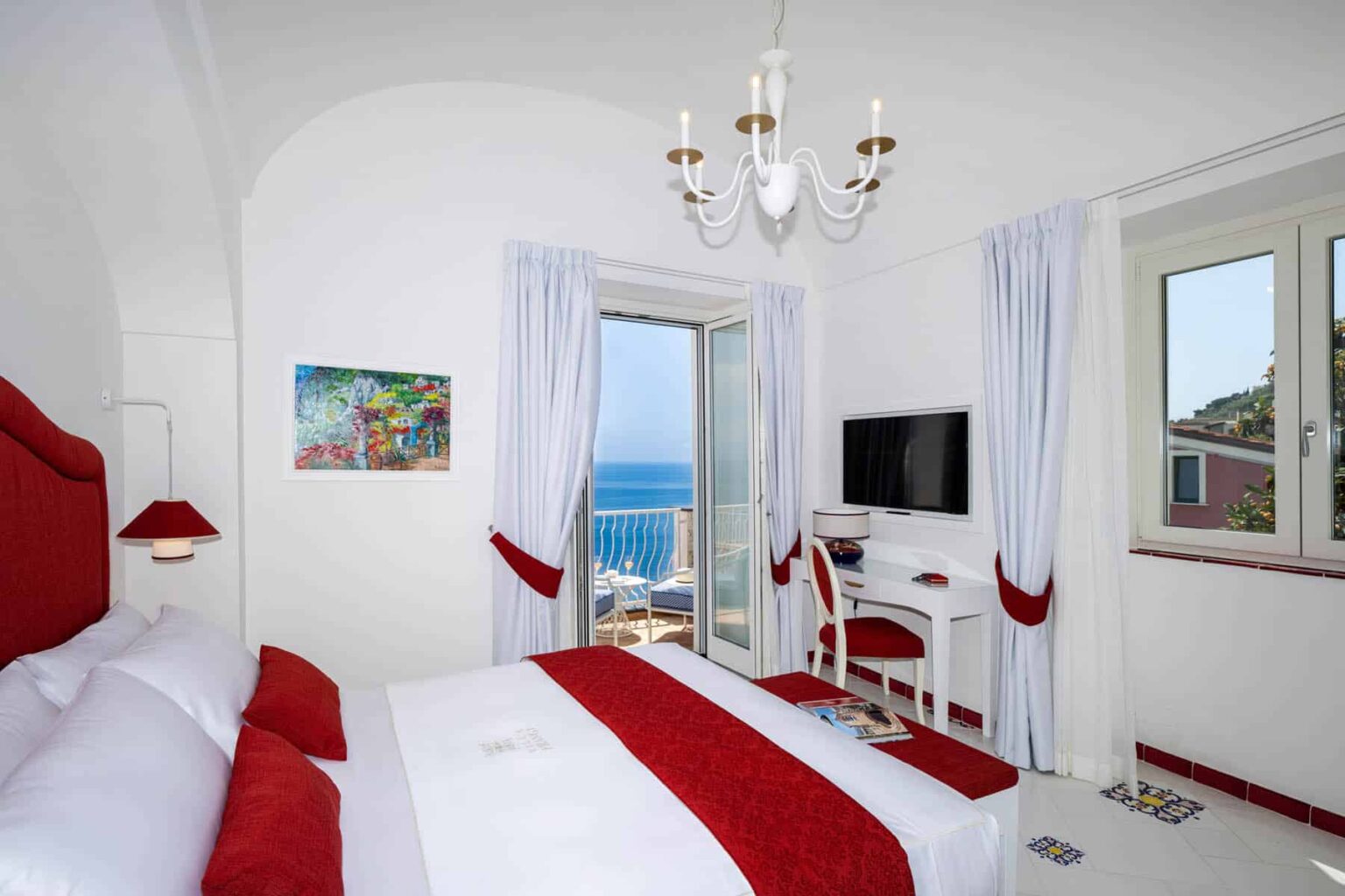 Bright and airy hotel room featuring a white and red color scheme, with a large window offering a view of the sea, a comfortable bed, a stylish chandelier, and a modern desk area. | Only on SopranoVillas