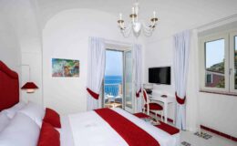 Bright and airy hotel room featuring a white and red color scheme, with a large window offering a view of the sea, a comfortable bed, a stylish chandelier, and a modern desk area.