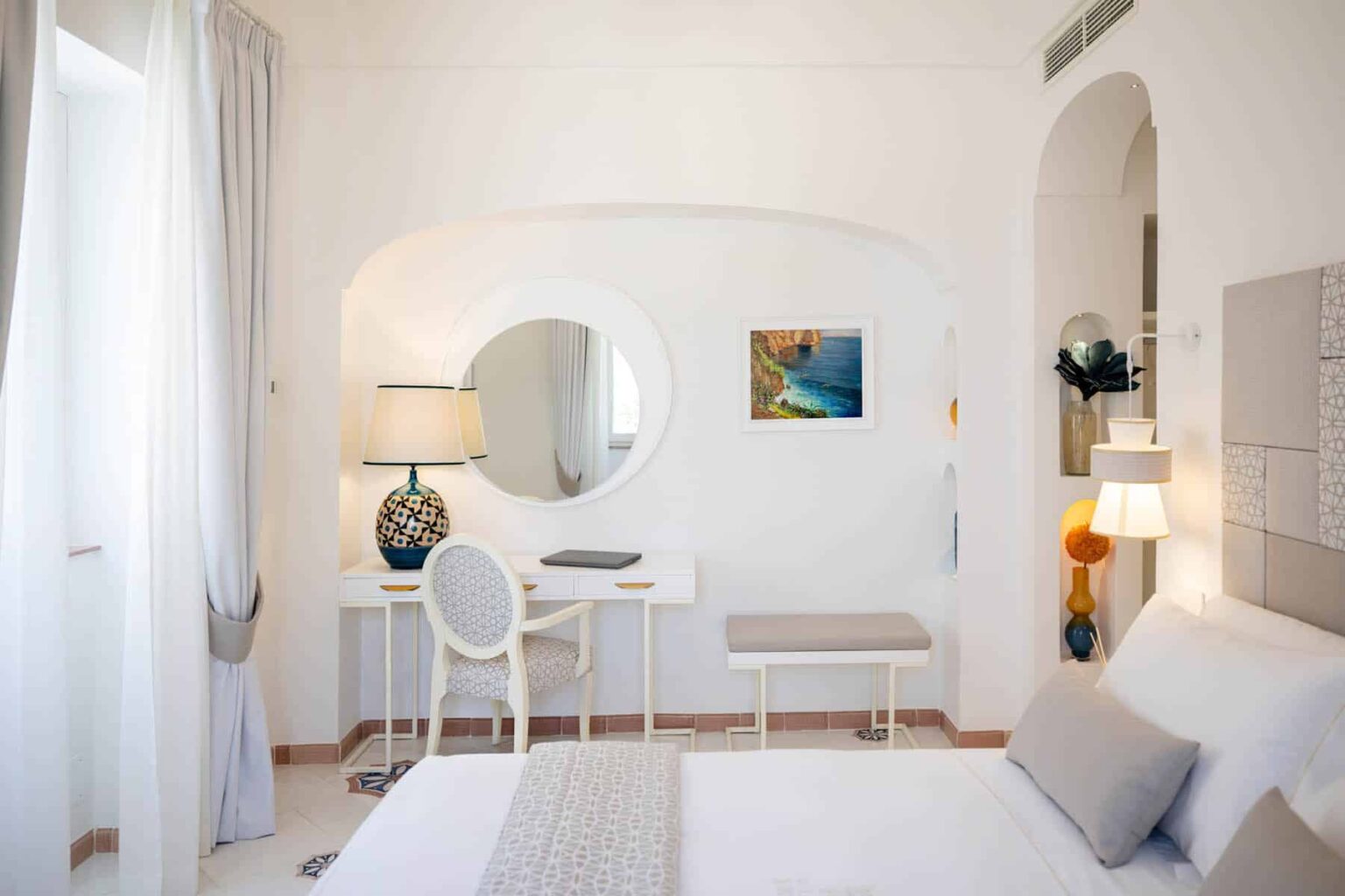 Bright and modern hotel room featuring a comfortable bed, stylish desk area with a lamp, and decorative artwork. The room has light-colored walls, large windows with sheer curtains, and contemporary furnishings, creating a welcoming atmosphere. | Only on SopranoVillas