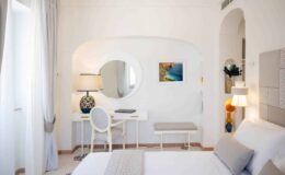 Bright and modern hotel room featuring a comfortable bed, stylish desk area with a lamp, and decorative artwork. The room has light-colored walls, large windows with sheer curtains, and contemporary furnishings, creating a welcoming atmosphere.