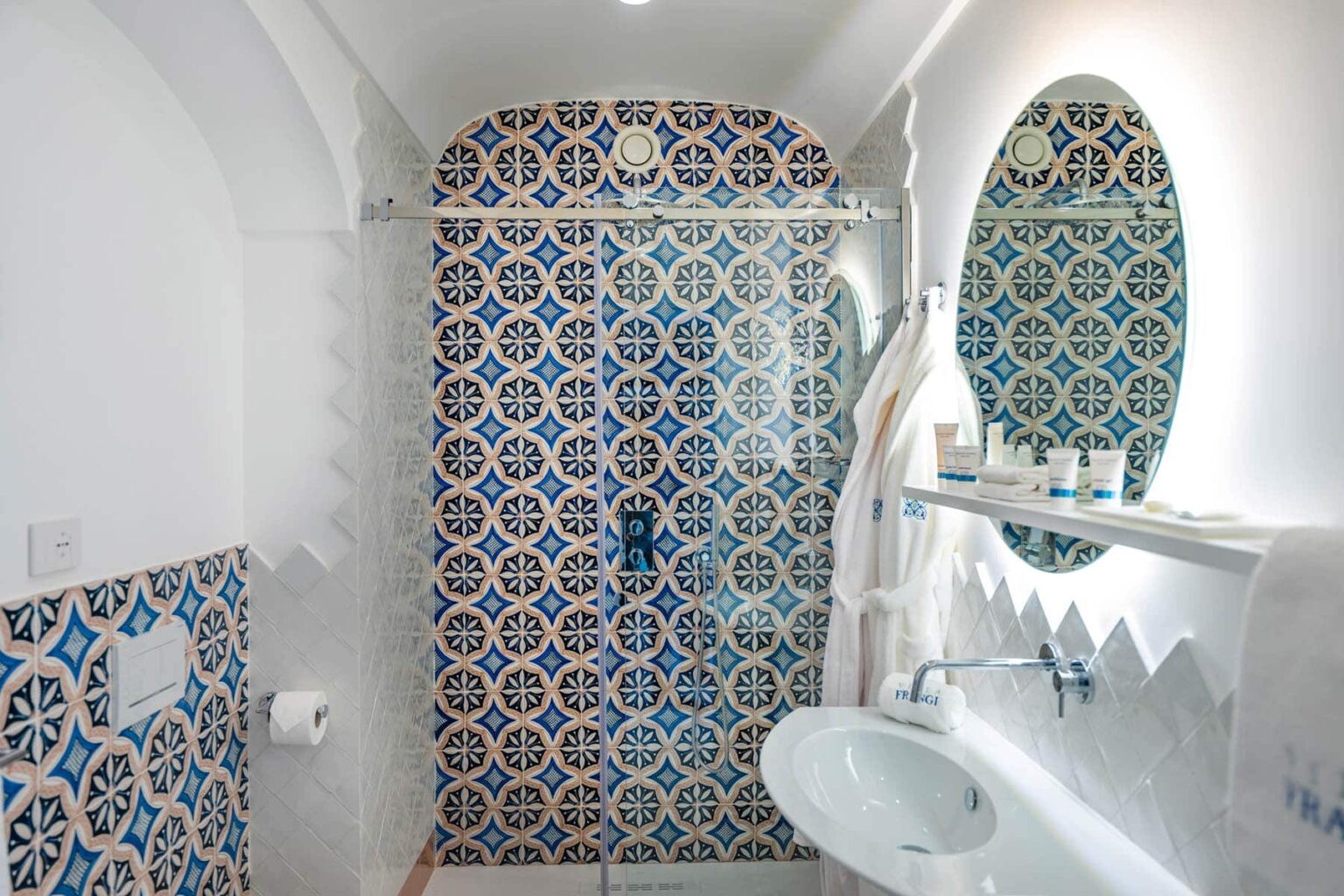 Bright and stylish bathroom featuring intricate blue and white patterned tiles, a glass shower enclosure, a modern sink, and a large oval mirror. The design combines elegance with functionality, creating a serene atmosphere. | Only on SopranoVillas