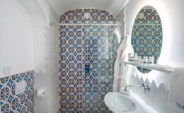Bright and stylish bathroom featuring intricate blue and white patterned tiles, a glass shower enclosure, a modern sink, and a large oval mirror. The design combines elegance with functionality, creating a serene atmosphere.