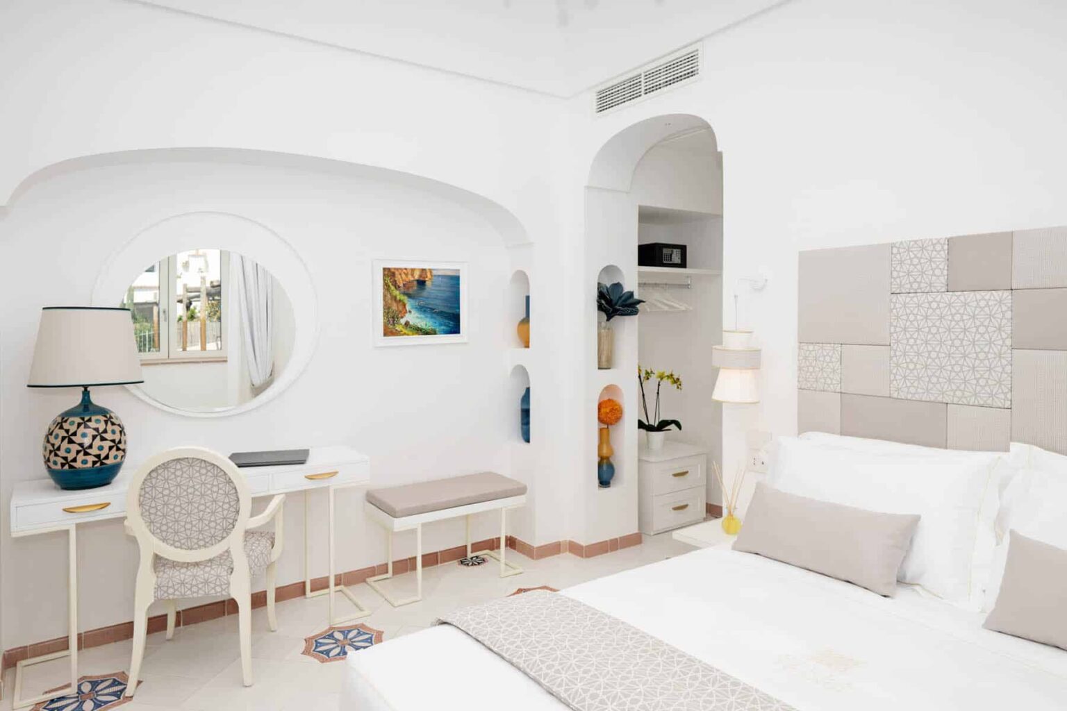Bright and modern hotel room featuring a white color scheme, a comfortable bed with neutral bedding, a stylish desk with a round mirror, and decorative artwork on the walls. The room is designed for relaxation, with natural light enhancing the inviting atmosphere. | Only on SopranoVillas