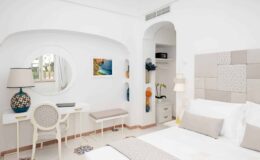 Bright and modern hotel room featuring a white color scheme, a comfortable bed with neutral bedding, a stylish desk with a round mirror, and decorative artwork on the walls. The room is designed for relaxation, with natural light enhancing the inviting atmosphere.