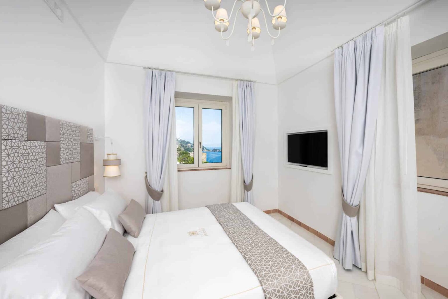 Modern hotel bedroom featuring a double bed, elegant light fixtures, and large windows with scenic views. The room is decorated in neutral tones with stylish curtains and a wall-mounted TV, creating a cozy and inviting atmosphere. | Only on SopranoVillas