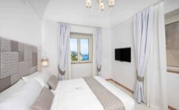 Modern hotel bedroom featuring a double bed, elegant light fixtures, and large windows with scenic views. The room is decorated in neutral tones with stylish curtains and a wall-mounted TV, creating a cozy and inviting atmosphere.