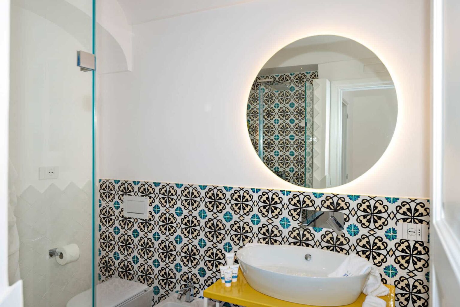 Modern bathroom featuring a stylish round mirror with ambient lighting, a sleek white sink, and vibrant patterned tiles in blue and black. The space includes a glass shower enclosure and a yellow accent table displaying toiletries. | Only on SopranoVillas