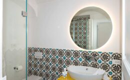Modern bathroom featuring a stylish round mirror with ambient lighting, a sleek white sink, and vibrant patterned tiles in blue and black. The space includes a glass shower enclosure and a yellow accent table displaying toiletries.