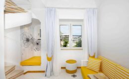 Bright and modern interior of a cozy room featuring white walls, yellow accents, a comfortable seating area with striped cushions, and a large window allowing natural light. The space includes a unique staircase and decorative elements, creating an inviting atmosphere.