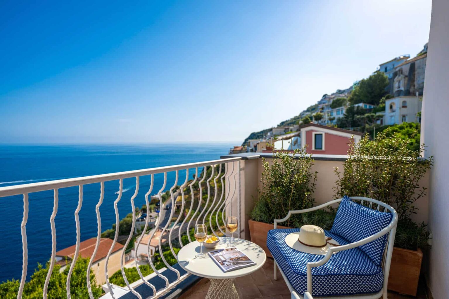Cozy balcony with a stunning sea view, featuring a small table with drinks and a sun hat, surrounded by lush greenery and colorful coastal homes in the background. Ideal for relaxation and enjoying the Mediterranean scenery. | Only on SopranoVillas