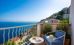 Cozy balcony with a stunning sea view, featuring a small table with drinks and a sun hat, surrounded by lush greenery and colorful coastal homes in the background. Ideal for relaxation and enjoying the Mediterranean scenery.