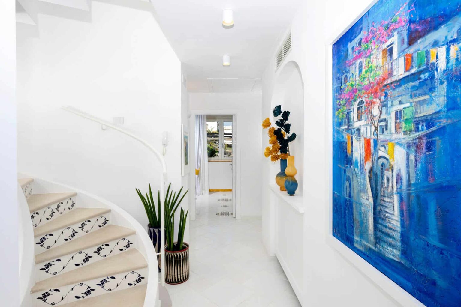 Bright and modern hallway featuring a spiral staircase, vibrant artwork on the wall, and decorative plants, creating an inviting atmosphere in a contemporary home. | Only on SopranoVillas