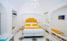 Bright and modern hotel bedroom featuring a white color scheme with yellow accents, a stylish shell-shaped headboard, elegant chandelier, and decorative black and white patterned flooring. Ideal for a luxurious stay.