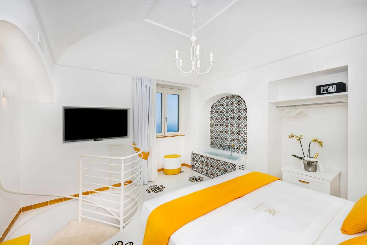 Bright and airy hotel room featuring white walls, a cozy bed with an orange accent, a flat-screen TV, and a window with scenic views. The room includes a stylish bathtub and modern decor, creating a relaxing atmosphere. | Only on SopranoVillas