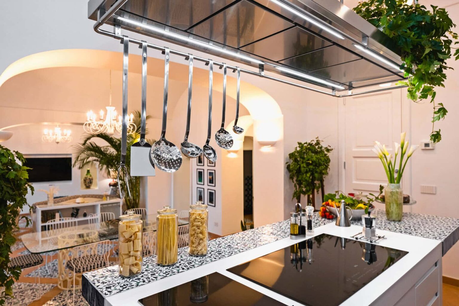 Modern kitchen interior featuring a sleek island with a cooktop, hanging ladles, and decorative pasta jars. Lush green plants and stylish lighting create a vibrant cooking space. | Only on SopranoVillas