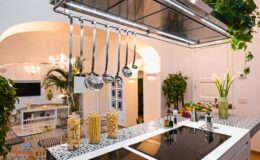 Modern kitchen interior featuring a sleek island with a cooktop, hanging ladles, and decorative pasta jars. Lush green plants and stylish lighting create a vibrant cooking space.