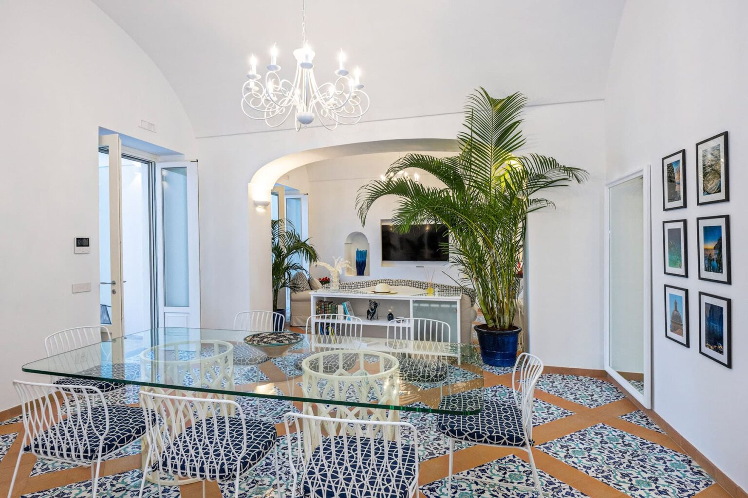 Bright and airy dining area featuring a glass table surrounded by white chairs, patterned tile flooring, and a large indoor plant. A modern chandelier hangs from the ceiling, while an open doorway leads to an outdoor space. The walls are adorned with framed photographs, and a cozy seating area is visible in the background. | Only on SopranoVillas