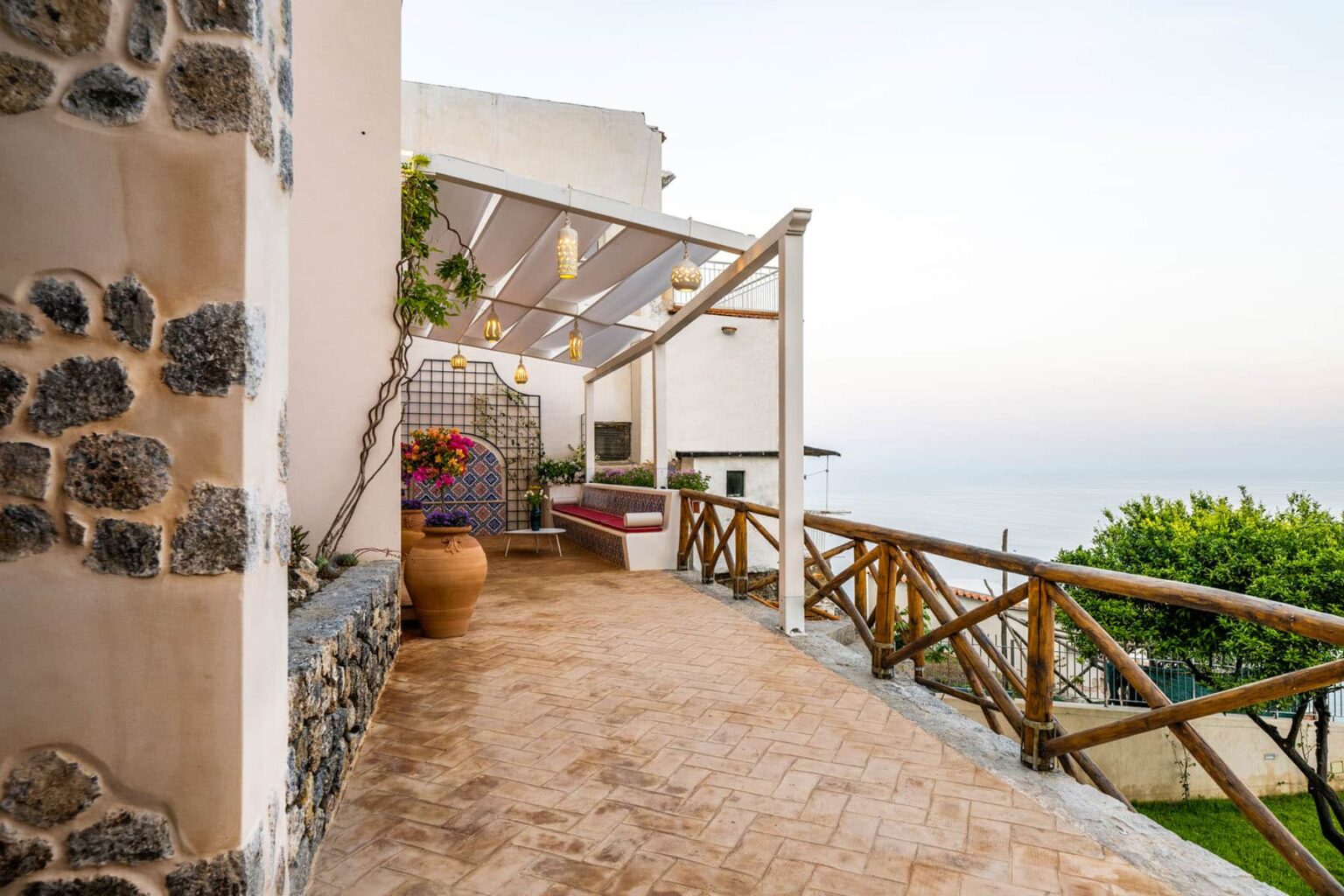 Scenic view of a coastal walkway with stone walls, wooden railing, and potted plants, leading to a tranquil ocean vista at sunset. | Only on SopranoVillas