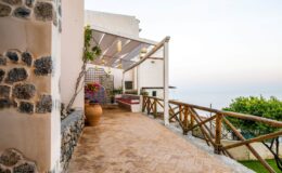 Scenic view of a coastal walkway with stone walls, wooden railing, and potted plants, leading to a tranquil ocean vista at sunset.