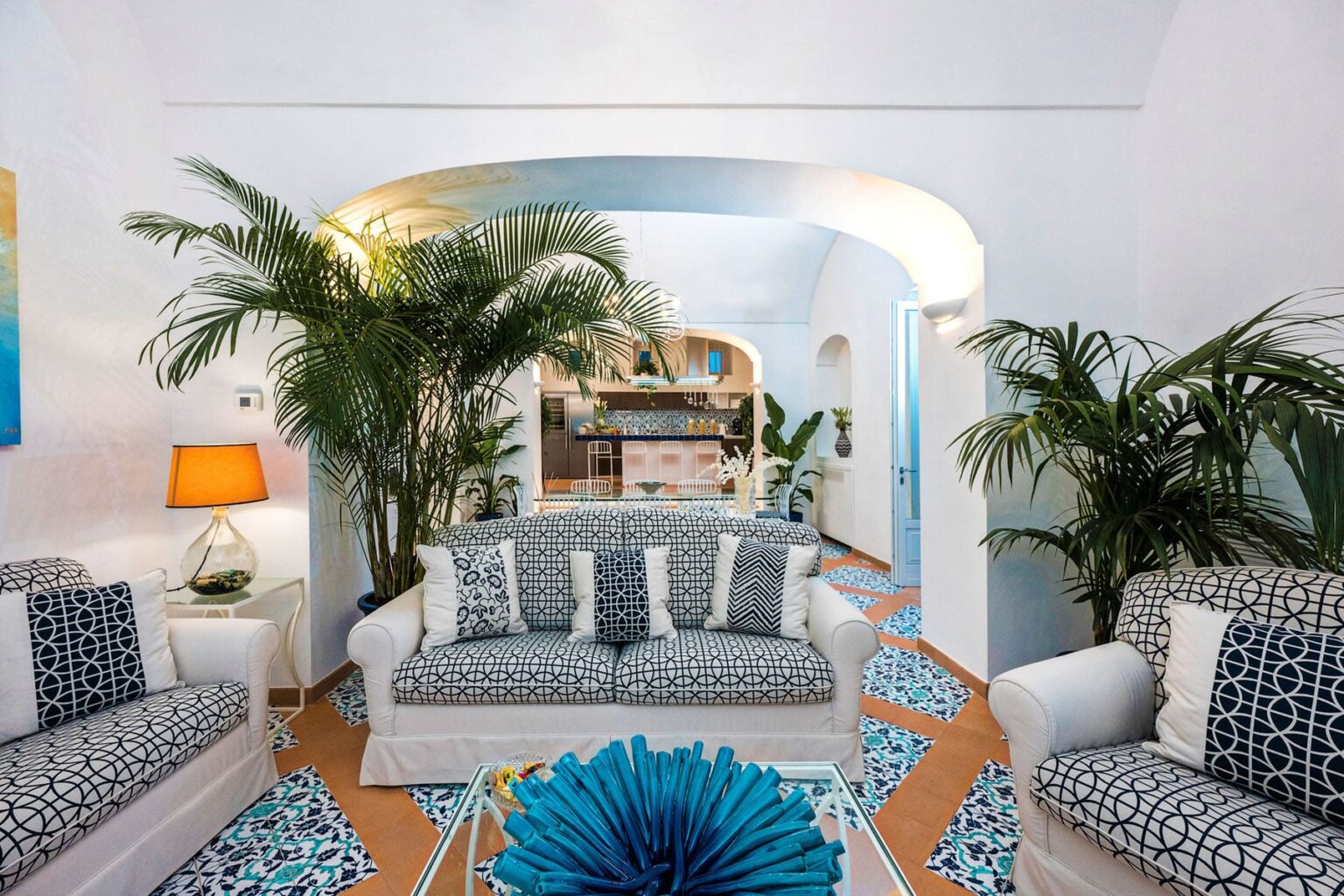Stylish living room featuring white sofas with blue accents, tropical plants, an artistic lamp, and vibrant patterned tiles, creating a bright and inviting atmosphere. | Only on SopranoVillas