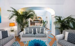 Stylish living room featuring white sofas with blue accents, tropical plants, an artistic lamp, and vibrant patterned tiles, creating a bright and inviting atmosphere.