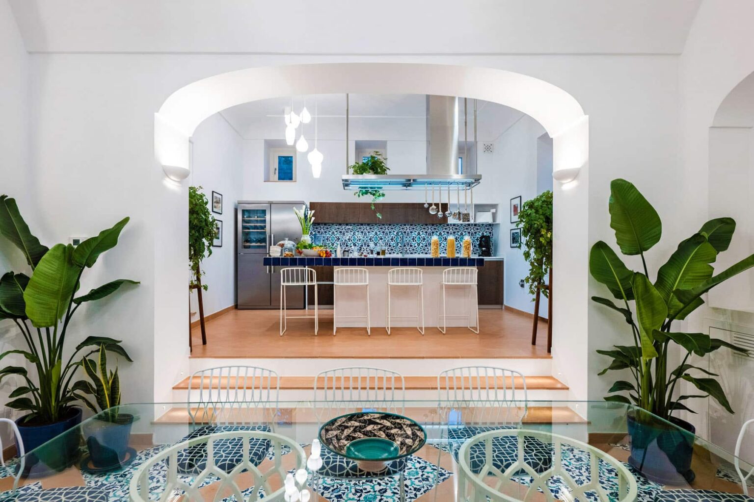 Modern kitchen design featuring a blue tiled backsplash, white bar stools, and indoor plants, creating a bright and inviting atmosphere. The open layout connects the kitchen to a dining area with stylish decor. | Only on SopranoVillas