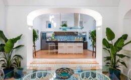 Modern kitchen design featuring a blue tiled backsplash, white bar stools, and indoor plants, creating a bright and inviting atmosphere. The open layout connects the kitchen to a dining area with stylish decor.
