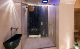 Modern shower design featuring glass doors, herringbone tile wall, wooden bucket for water, and stylish sink. The space is illuminated with soft lighting, creating a luxurious and relaxing atmosphere.