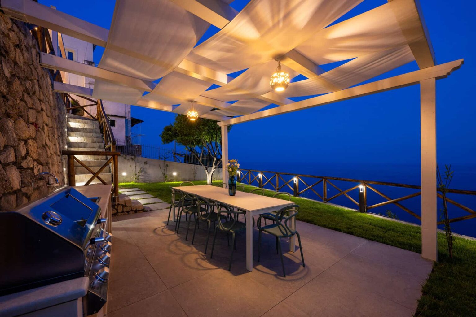 Outdoor dining area with a modern table and chairs under a stylish pergola, featuring ambient lighting and a scenic view at dusk. A barbecue grill is positioned nearby, enhancing the outdoor entertaining space. | Only on SopranoVillas