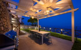 Outdoor dining area with a modern table and chairs under a stylish pergola, featuring ambient lighting and a scenic view at dusk. A barbecue grill is positioned nearby, enhancing the outdoor entertaining space.