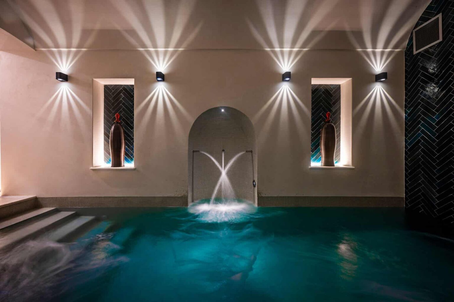 Indoor spa area featuring a tranquil pool with water features, illuminated by stylish wall sconces and ambient lighting, creating a serene atmosphere for relaxation and wellness. | Only on SopranoVillas