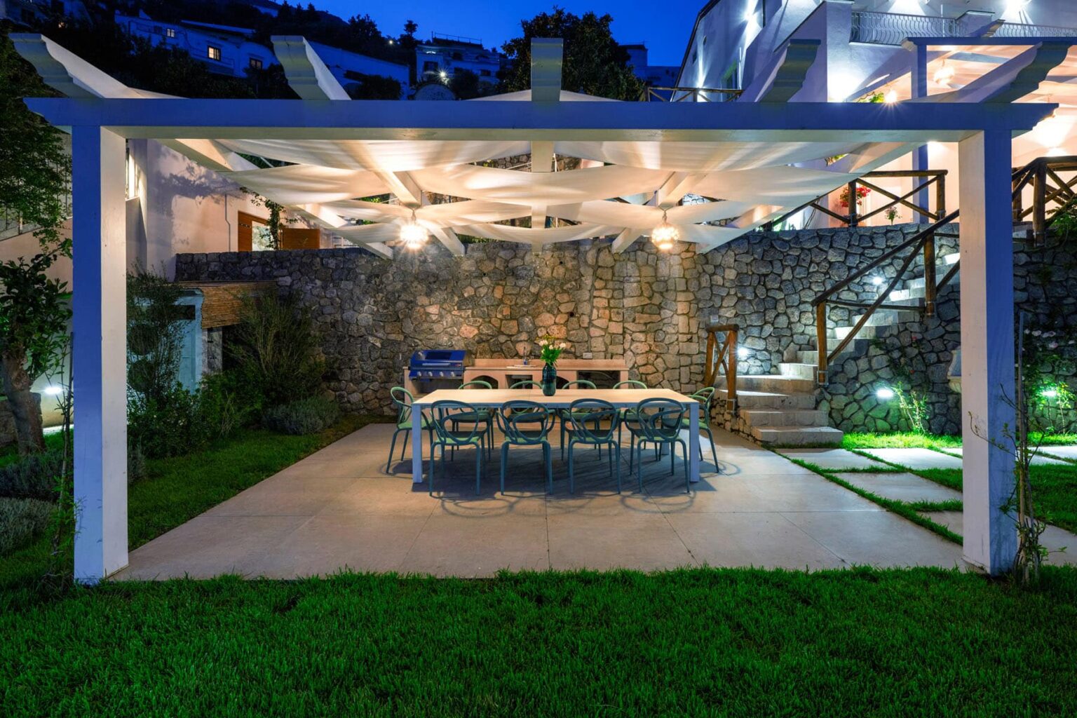 Outdoor dining area featuring a large table and chairs under a pergola, illuminated by soft lighting, surrounded by lush greenery and stone walls, creating a cozy atmosphere for evening gatherings. | Only on SopranoVillas