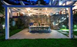 Outdoor dining area featuring a large table and chairs under a pergola, illuminated by soft lighting, surrounded by lush greenery and stone walls, creating a cozy atmosphere for evening gatherings.
