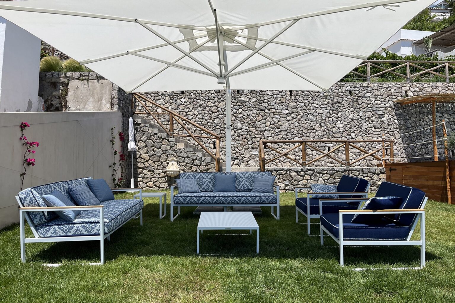 Outdoor seating area featuring a stylish arrangement of blue and white patio furniture under a large umbrella, set on green grass with a stone wall backdrop. Ideal for relaxation and entertaining. | Only on SopranoVillas
