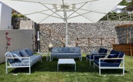 Outdoor seating area featuring a stylish arrangement of blue and white patio furniture under a large umbrella, set on green grass with a stone wall backdrop. Ideal for relaxation and entertaining.