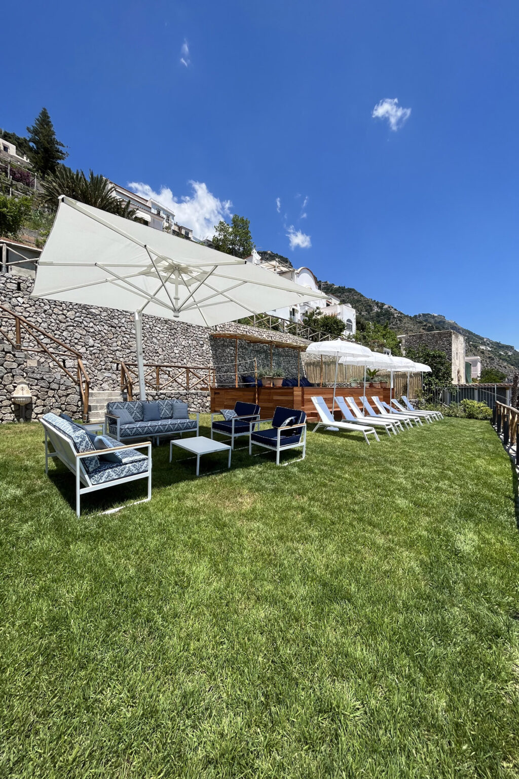 Lounge area with comfortable seating and sun loungers under umbrellas, surrounded by lush green grass and stone walls, ideal for relaxation in a sunny outdoor setting. | Only on SopranoVillas