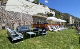 Lounge area with comfortable seating and sun loungers under umbrellas, surrounded by lush green grass and stone walls, ideal for relaxation in a sunny outdoor setting.