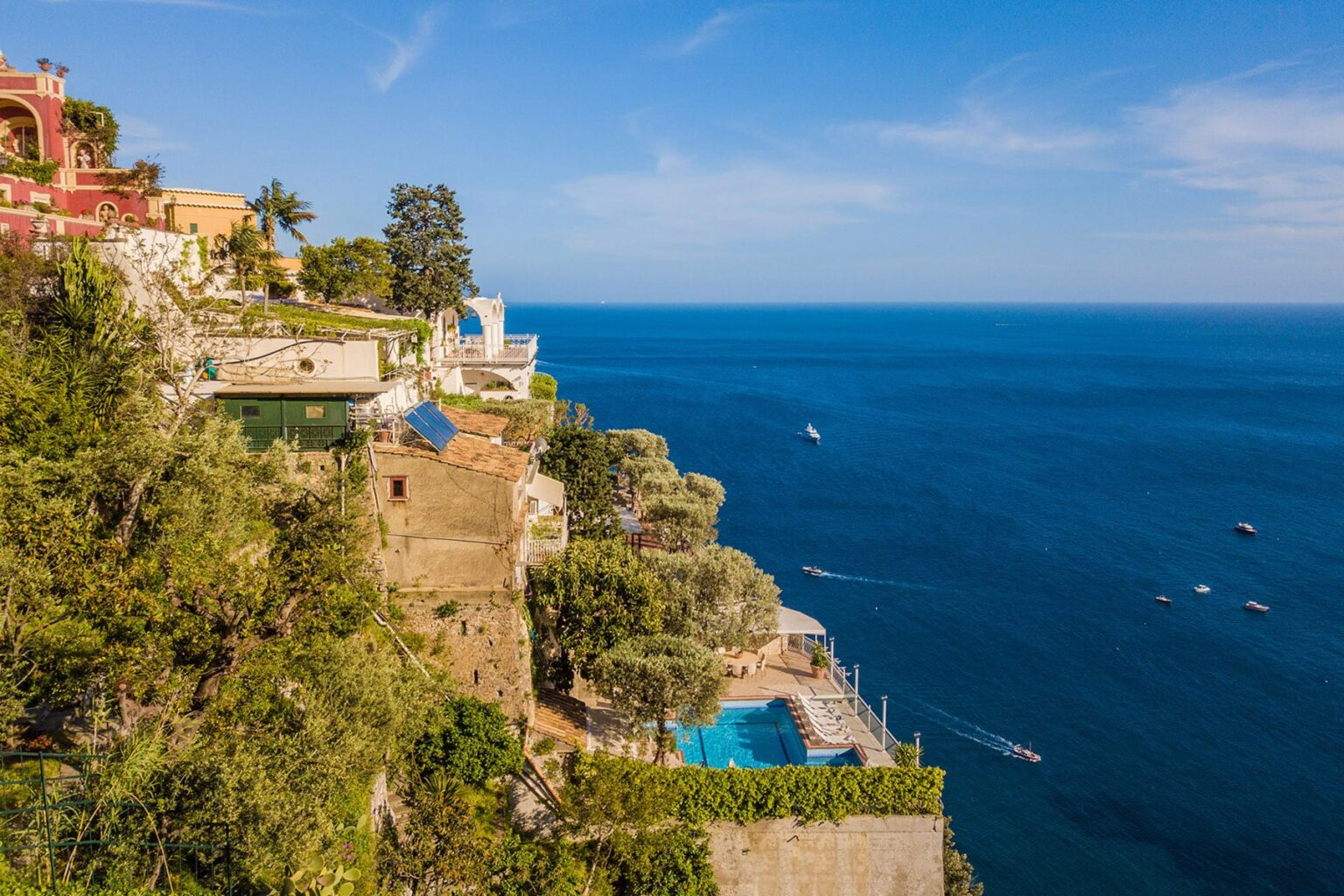 Stunning coastal view of a cliffside villa overlooking the blue sea, with a swimming pool and lush greenery along the shoreline, highlighting the beauty of a Mediterranean landscape. | Only on SopranoVillas