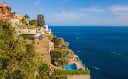 Stunning coastal view of a cliffside villa overlooking the blue sea, with a swimming pool and lush greenery along the shoreline, highlighting the beauty of a Mediterranean landscape.