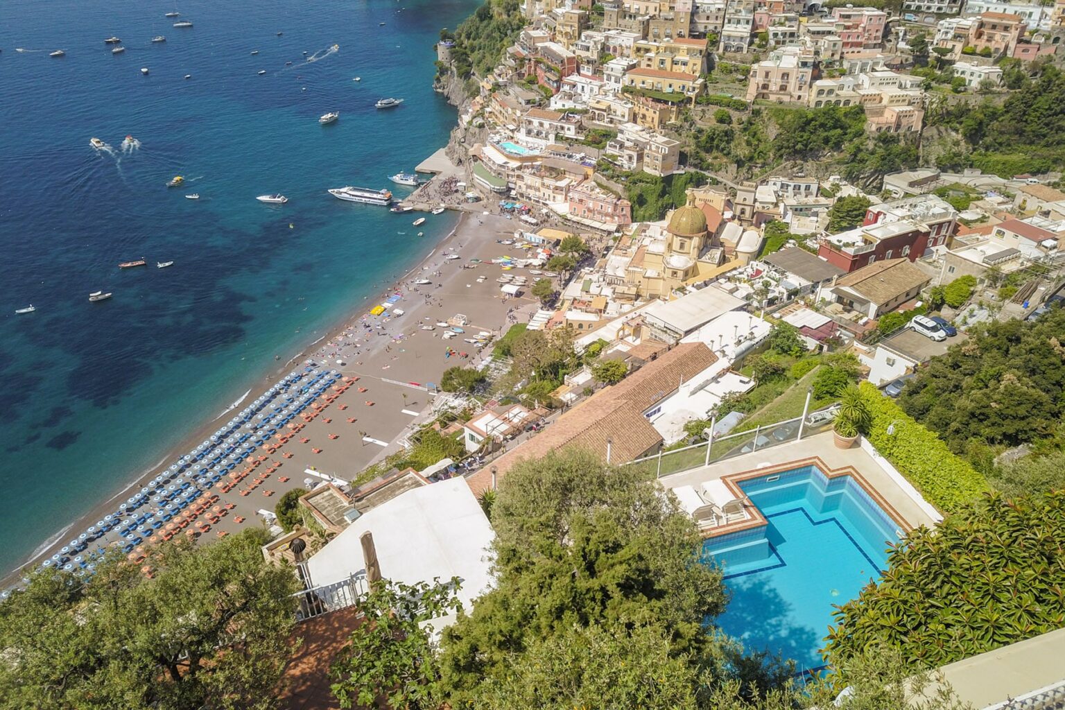 Aerial view of Positano, Italy, showcasing the beach, colorful hillside buildings, and a swimming pool in the foreground, with boats in the turquoise sea. | Only on SopranoVillas